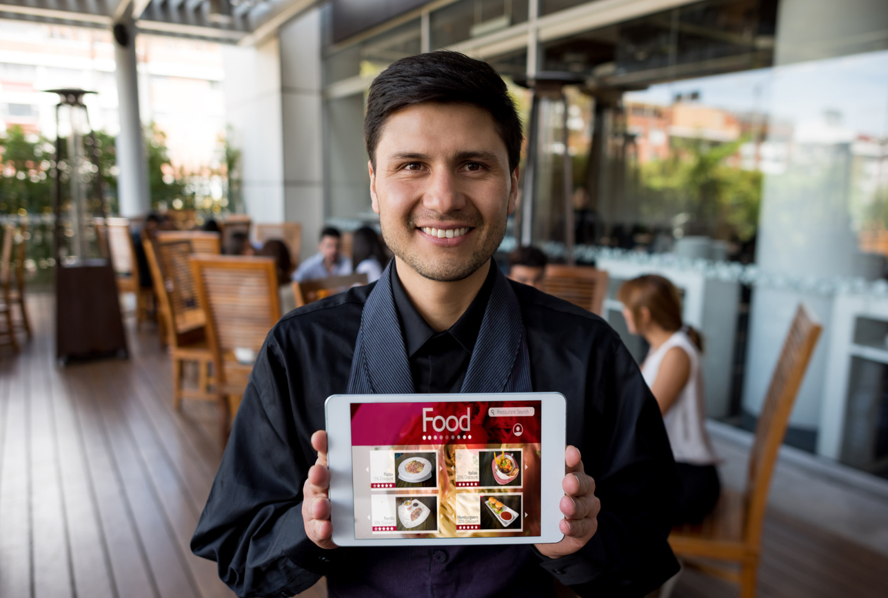 Friendly waiter holding a digital tablet showing the menu looking at camera smiling
